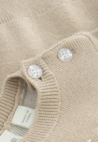 Pull en tricot beige avec un col rond, arborant deux boutons blancs. Le tissu présente une texture lisse avec un bord côtelé. L'étiquette indique "Fabriqué en Chine."