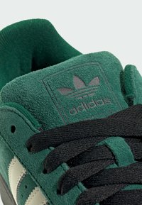 Groene suède sneaker met zwarte veters, voorzien van een gestructureerde tong met een zwart adidas-logo en drie witte strepen aan de zijkant.