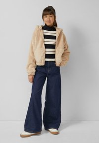 Beige, pluche gewatteerde jas met capuchon over een zwart-wit gestreepte, geribbelde trui, gecombineerd met wijde donkerblauwe jeans en witte sneakers.