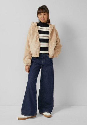 Beżowa puchata kurtka z kapturem na czarno-białym prążkowanym, prążkowanym swetrze, w połączeniu z szerokimi, ciemnoniebieskimi jeansami i białymi sneakersami.