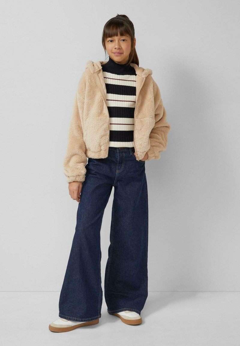 Beige, pluche gewatteerde jas met capuchon over een zwart-wit gestreepte, geribbelde trui, gecombineerd met wijde donkerblauwe jeans en witte sneakers.