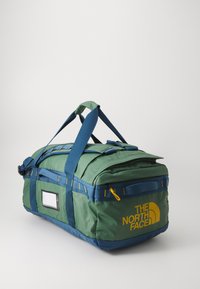Grønn og blå duffelbag med gule detaljer, laget av slitesterkt stoff. Har to håndtak, en skulderreim og et frontområde for etikett.