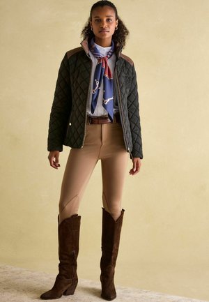 Schwarze gesteppte Jacke mit braunen Schulterakzenten, kombiniert mit beigen, figurschmeichelnden Leggings und braunen Wildleder-Stiefeln bis zum Knie. Blauer gemusterter Schal inklusive.