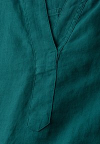 Türkisfarbene Leinenstoffe mit einem Ausschnitt und gesteppten Details. Weiche Textur mit einem leichten Glanz, die ein sauberes, minimalistisches Design zur Schau stellt.