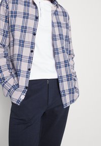 Ruudullinen flanellipaita vaaleanharmaana, jossa on sinisiä ja laivastonsinisiä raitoja, kerrostettuna valkoisen henley-paidan päälle, yhdistettynä tummiin kuvioituihin housuihin.