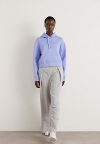 Haut à capuche cropped violet clair avec cordon de serrage et poignets côtelés, associé à un pantalon de survêtement large gris et des baskets blanches. Fond uni.