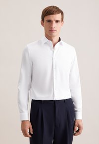 Camicia bianca a maniche lunghe con colletto appuntito, chiusura frontale con bottoni e texture liscia. Abbinata a pantaloni sartoriali blu scuro.