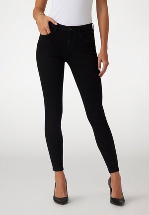 Femme portant un jean skinny noir et des talons hauts noirs, une main dans la poche et un haut blanc sans manches.