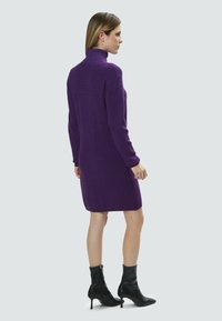 Robe en tricot violette à col roulé avec des manches longues, ourlet à hauteur des genoux et texture côtelée, associée à des bottines noires. 