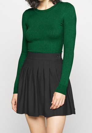 Mujer con un top ajustado de manga larga verde acanalado, metido dentro de una minifalda plisada negra de talle alto con detalle de botones.