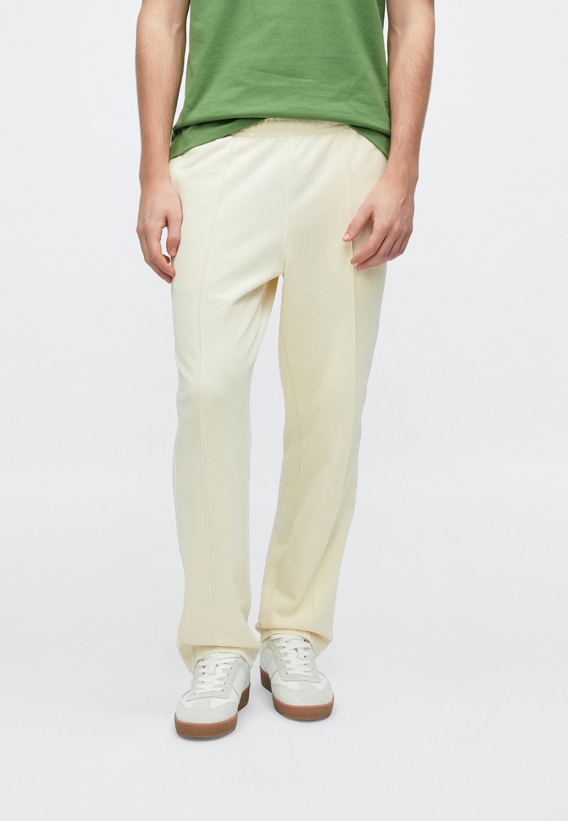 Παντελόνια σε κρεμ απόχρωση με άνετη εφαρμογή, με λεία υφή και διακριτικές πιέτες στο μπροστινό μέρος, συνδυασμένα με λευκά και γκρι αθλητικά παπούτσια.