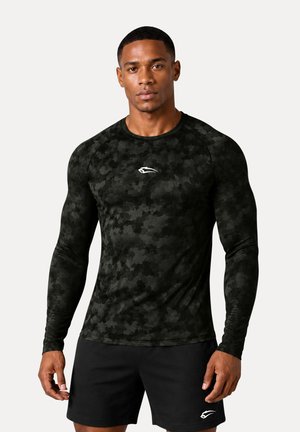 Muskuløs mand iført en langærmet sort camouflage kompressionsshirt og sorte atletikshorts med hvidt logo på hvert stykke tøj.