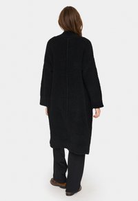 Svart långärmad kappa gjord av texturerat tyg, med en avslappnad passform, hög krage och rak fåll. Synliga sömmar på baksidan.