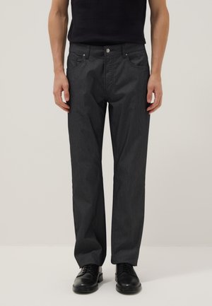 Hombre vestido con pantalones negros de pierna recta, zapatos negros y camisa negra, de pie contra un fondo claro y liso.