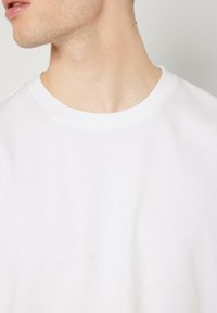 Balta kokvilnas T-kreklu ar klasisku apaļu kakla izgriezumu, īsām piedurknēm un gludu tekstūru, izceļot tīru un minimālistisku dizainu.