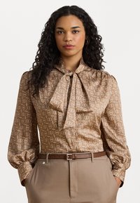 Brun mønstrete bluse med sløyfe, lange puffede ermer og glatt tekstur, kombinert med beige høyde liv bukser og et brunt belte.