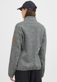 Graue Fleecejacke mit hohem Kragen, strukturiertem Stoff und einem Lederlogo-Patch am linken Arm. Kombiniert mit einer schwarzen Kappe.