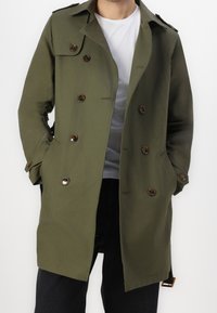 Homme portant un trench-coat olive à double boutonnage avec des boutons marron et les mains dans les poches, par-dessus un t-shirt blanc et un pantalon noir.