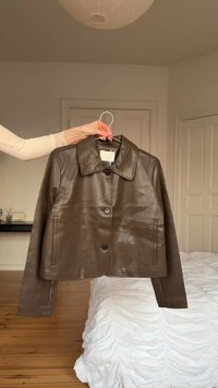 Χέρι που κρατά ένα σκούρο καφέ δερμάτινο μπουφάν σε κρεμάστρα σε υπνοδωμάτιο με ξύλινο πάτωμα και λευκό κάλυμμα κρεβατιού.