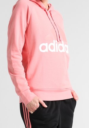 Henkilö pukeutuneena vaaleanpunaiseen Adidas-huppariin, jossa valkoinen logo, sekä mustiin housuihin, joissa vaaleanpunaiset raidat, kädet taskuissa, seisoo yksinkertaisen taustan edessä.