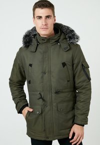 Parka verde olivo con capucha forrada de piel, cierres de cremallera y a presión, cuatro bolsillos frontales y cordones ajustables en la cintura.