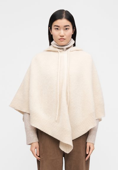 Poncho en laine beige avec capuche, présentant une texture lisse et des accents côtelés horizontaux le long de l'ourlet, porté par-dessus un pull léger à col roulé.