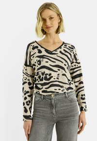 Suéter beige con estampado animal negro, escote en V y mangas largas, combinado con jeans grises. El diseño presenta patrones y texturas variadas.