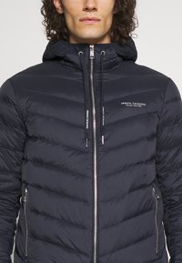 Marineblaue Pufferjacke mit Reißverschluss and vorne, Seitentaschen und einer verstellbaren Kapuze. Verfügt über ein gestepptes Muster und Logodetail auf der Brust.