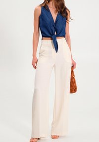 Haut sans manches en denim bleu avec n œud à l'avant, associé à un pantalon large crème. Sac à main tressé marron ajoutant de la texture et du contraste.