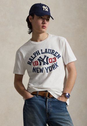 T-shirt en coton blanc avec un imprimé bleu et rouge "Ralph Lauren 1903 New York". Assorti à un jean en denim bleu et une casquette marine.