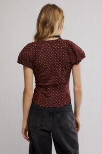 Μπορντό κοντομάνικο τοπ με χρυσές πουά, εφαρμοστή γραμμή και βολάν στη λαιμόκοψη. Συνδυάζεται με ψηλόμεσα μαύρα τζιν, που δείχνουν λεπτομέρειες στην πλάτη.