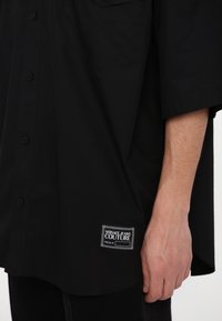 Black short-sleeve button-up shirt with a relaxed fit, featuring a visible label on the left side and a smooth finish.