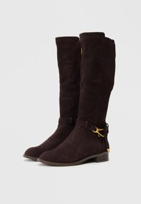Čizme od tamnosmeđeg sueda do koljena s glatkom teksturom, remenom oko gležnja sa zlatnim detaljem i niskim, složenim potplatom.