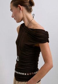 Mujer con una blusa negra, de tirantes caídos y fruncida, que lleva un cinturón con tachuelas plateadas, y un collar largo negro con un colgante ovalado plateado.
