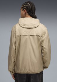 Beige vindjakke med hætte, med et bagventil og elastiske manchetter. Glat, let materiale, designet til komfort og bevægelighed.