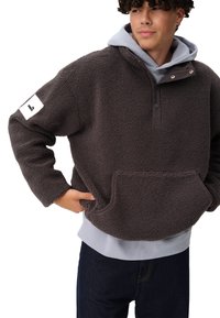 Polarowy pulower w ciemnoszarym kolorze, z wysokim kołnierzem z guzikami, kieszenią z przodu oraz białą metką na lewym rękawie. Teksturowana powierzchnia.