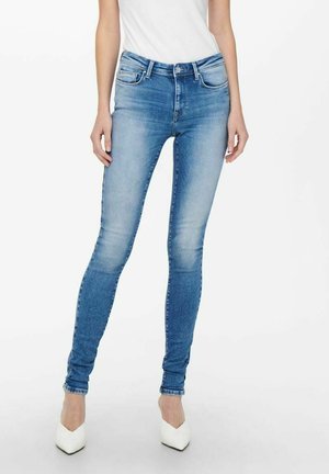 Vaqueros ajustados en denim azul claro, con un diseño clásico de cinco bolsillos, detalles desgastados y pierna ajustada. Llevados con tacones blancos.