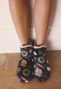 Fuzzy, navy slippers with a holiday pattern featuring gingerbread houses, snowmen, and wreaths. Soft, fur-lined cuff for added comfort.