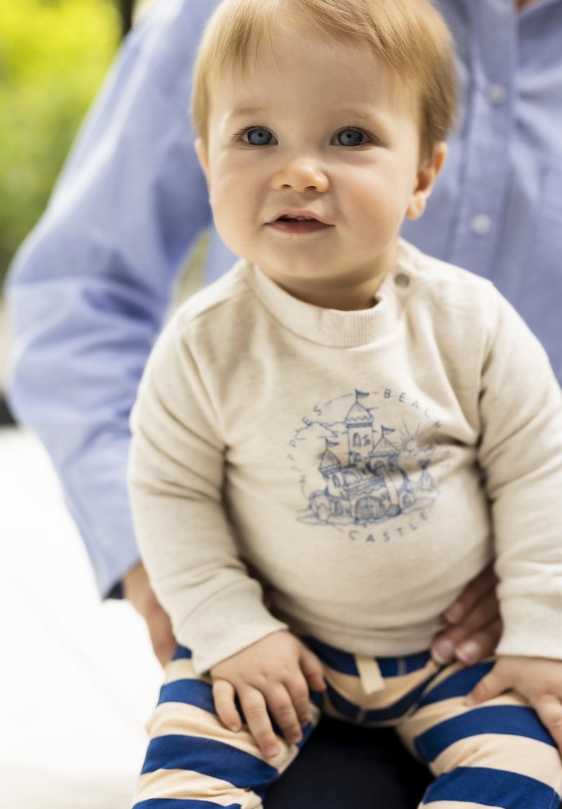 Chemise beige clair à manches longues avec un motif de château bleu, associée à des leggings rayés bleu marine et beige. Les mains potelées de l'enfant sont visibles.