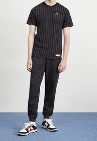 Persona vistiendo una camiseta negra de rayas de Jordan, pantalones jogger negros y zapatillas Nike blancas, negras y rojas, de pie sobre una alfombra azul.