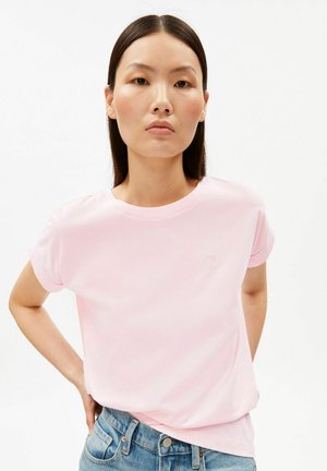 Junge Frau mit glatten dunklen Haaren, die ein hellrosa T-Shirt und blaue Jeans trägt, steht mit einer Hand in der Hüfte vor weißem Hintergrund.