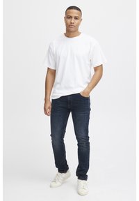 Vit bomull T-shirt med kort ärm, i kombination med mörkblå smala jeans och vita sneakers, stående mot en ljus bakgrund.
