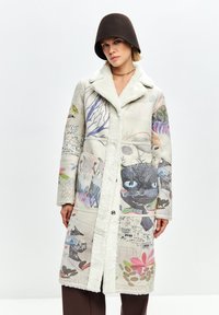 Donna che indossa un lungo cappotto in shearling color crema con illustrazioni colorate di gatti e fiori, abbinato a pantaloni marroni e un cappello cloche marrone scuro.