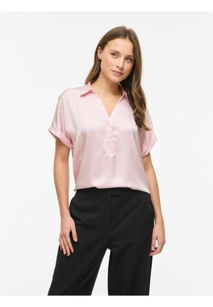 Kvinne iført en lys rosa kortermet knappet bluse og svarte bukser, stående mot en ensfarget hvit bakgrunn.