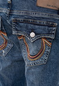 Bolsillo trasero de jeans de mezclilla con solapa, cierre de botón, costuras curvas multicolores y parche de cuero marrón con la etiqueta "True Religion".