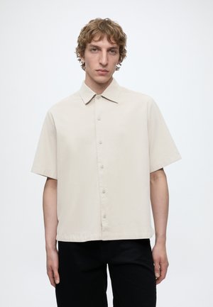 Jeune homme aux cheveux ondulés portant une chemise beige clair à manches courtes boutonnée et un pantalon noir, debout devant un fond uni.
