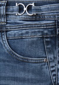 Jean en denim bleu avec une boucle de ceinture circulaire en argent, des coutures contrastées et une poche avant ornée d'un accent métallique circulaire.