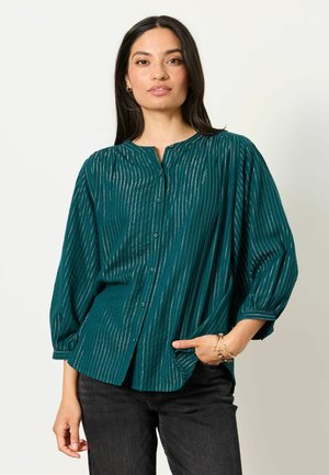 Young woman with long dark hair wearing a teal button-up blouse with metallic vertical stripes and black pants, hand in pocket, neutral background.
