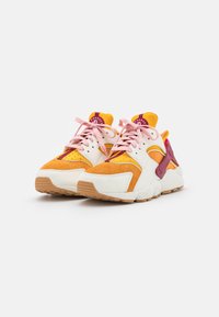 Ett par färgglada träningsskor med orange, vita och bourgogne paneler, rosa snören och texturerade bruna sulor på en vit bakgrund.