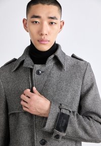 Grå, texturerad överrock med hög krage, knappstängning framtill och justerbara ärmslut. Har en etikett på ärmen och ett slätt innerfoder.